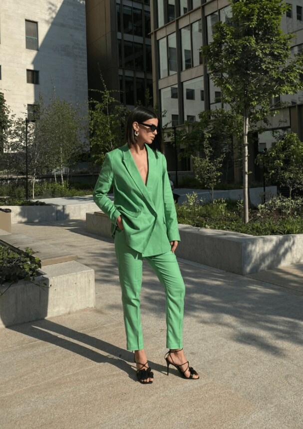 Un model într-un costum verde, tocuri înalte negre și ochelari de soare stilați pe fundalul unui oraș însorit
