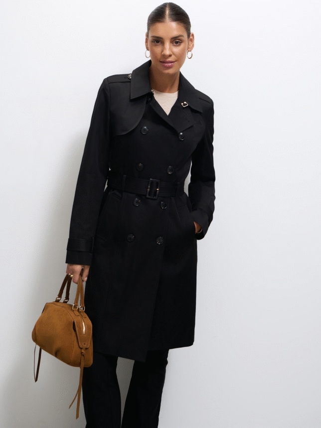 O femeie într-un elegant trench negru, cu două rânduri de nasturi, și curea în talie. Modelul ține o geantă maro; are părul legat și poartă cercei rotunzi aurii.