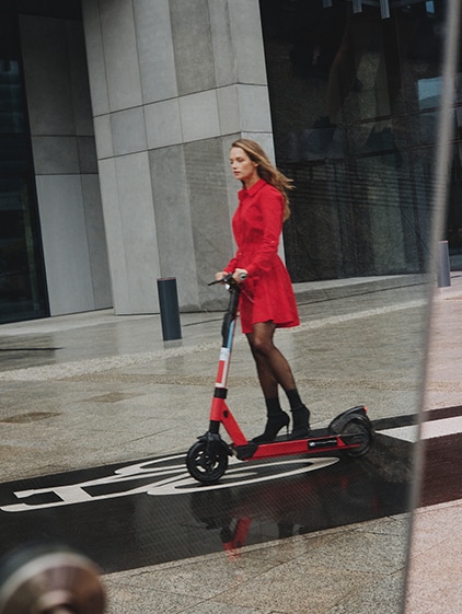 o femeie care merge pe un scuter electric, într-o rochie roșie și cizme cu toc înalt
