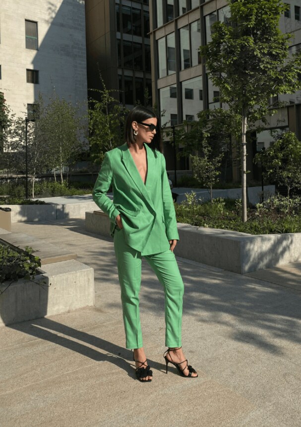 Un model într-un costum verde, tocuri înalte negre și ochelari de soare stilați pe fundalul unui oraș însorit

