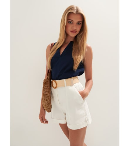 un model într-o ținută smart casual de vară: o cămașă bleumarin fără mâneci și pantaloni scurți albi cu curea
