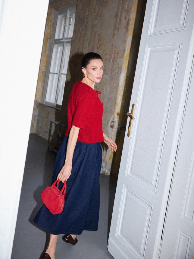 Modelka w walentynkowej stylizacji: czerwony krótki kardigan z wiązaniami na kokardki, szeroka jeansowa spódnica midi i mała czerwona torebka do ręki.