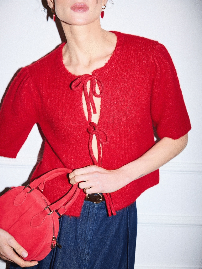 Romantyczna stylizacja na randkę 2026: czerwony sweter z krótkim rękawem typu "coquette" z kokardami, ciemna spódnica i zamszowa torebka w kolorze intensywnej czerwieni.