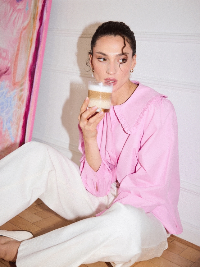 Kobieta w różowej koszuli z dużym kołnierzem z falbanką i białych spodniach, siedząca na podłodze z kawą latte. Inspiracja na walentynkowy poranek.