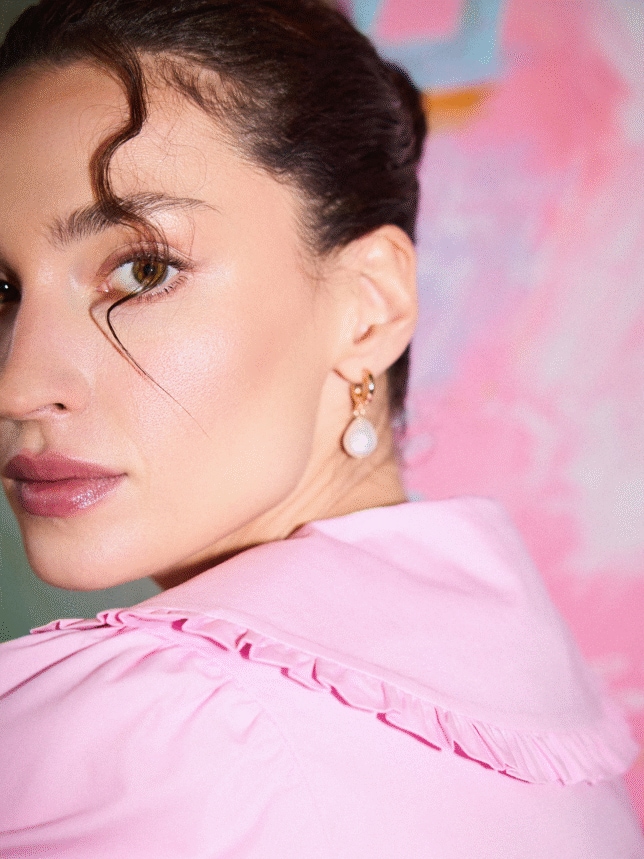 Zbliżenie na twarz kobiety w delikatnym makijażu typu "clean girl", kolczyki z perłami i różową bluzkę z ozdobnym kołnierzem na tle abstrakcyjnego różowego obrazu.