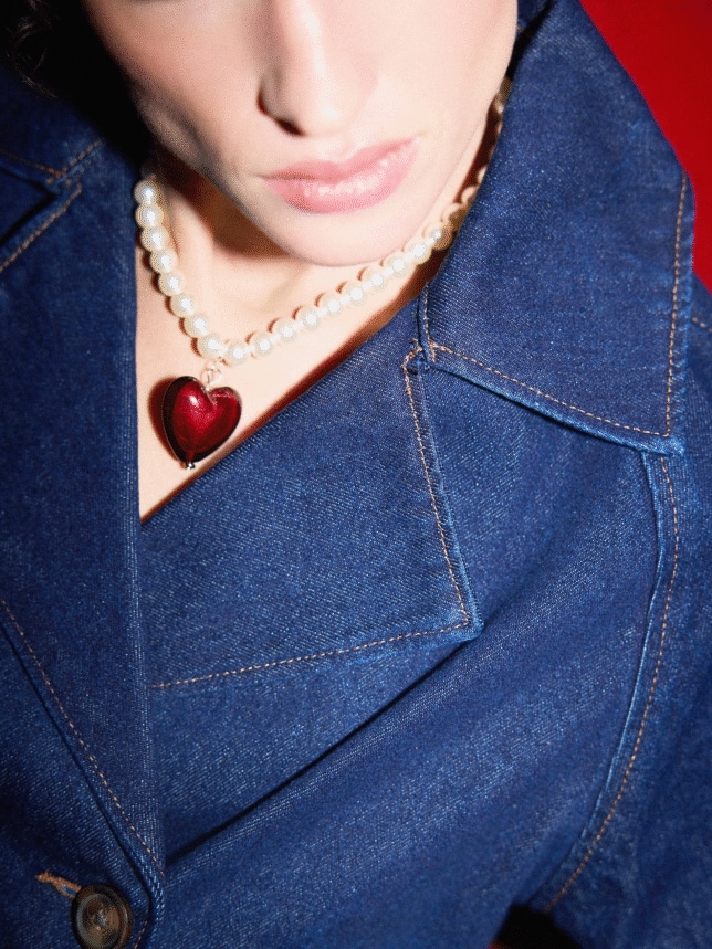 Zbliżenie na naszyjnik z białych pereł z dużą zawieszką w kształcie czerwonego serca, noszony do ciemnoniebieskiej kurtki z denimu.