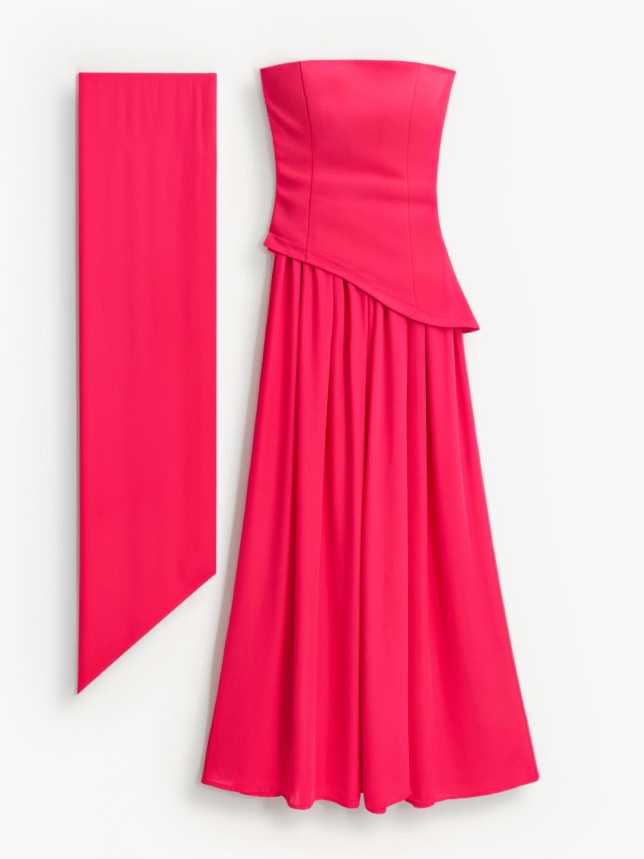 Długa suknia wieczorowa na wesele w intensywnym kolorze fuksji. Model z gorsetową górą bez ramiączek, asymetrycznym odcięciem w talii i szerokim, lejącym dołem. Obok widoczny długi, pasujący szal.