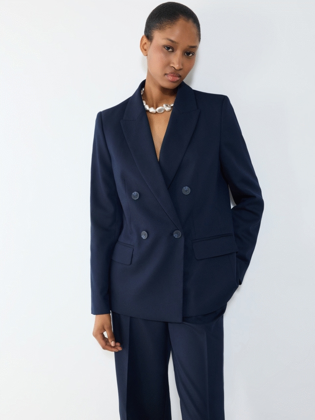 Portret kobiety w ciemnogranatowej marynarce o klasycznym kroju z podwójnym rzędem guzików i perłowym naszyjnikiem typu choker, na tle białej ściany.