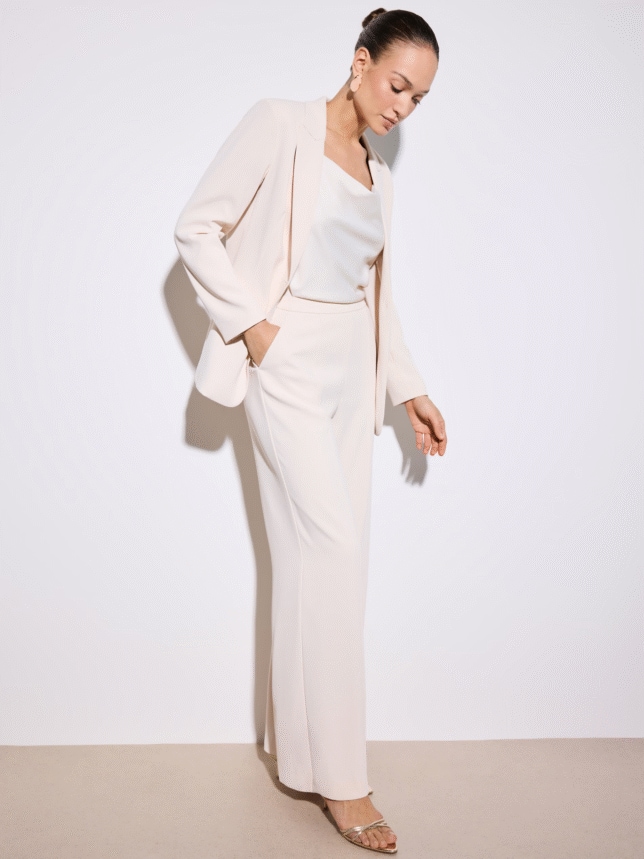 Kobieta w jasnym garniturze i połyskującym topie, prezentująca luźniejszy krój marynarki i eleganckie spodnie z kieszeniami.