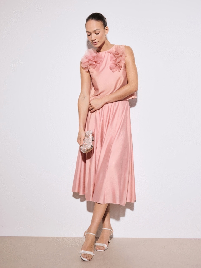 Wizytowy komplet damski w kolorze różowym: satynowy top z trójwymiarowymi kwiatami na ramionach oraz plisowana spódnica midi, zestawione z błyszczącą kopertówką.