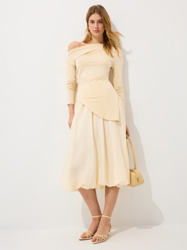 Elegancki damski komplet w kolorze waniliowym: asymetryczny top z długim rękawem odsłaniający jedno ramię oraz rozkloszowana spódnica midi.