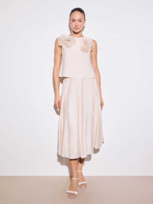 Stylizacja na wesele: jasny, pastelowy komplet składający się z luźnego topu z dekoracyjnymi kwiatami na ramionach oraz rozkloszowanej spódnicy, zestawiony z białymi sandałkami na obcasie.