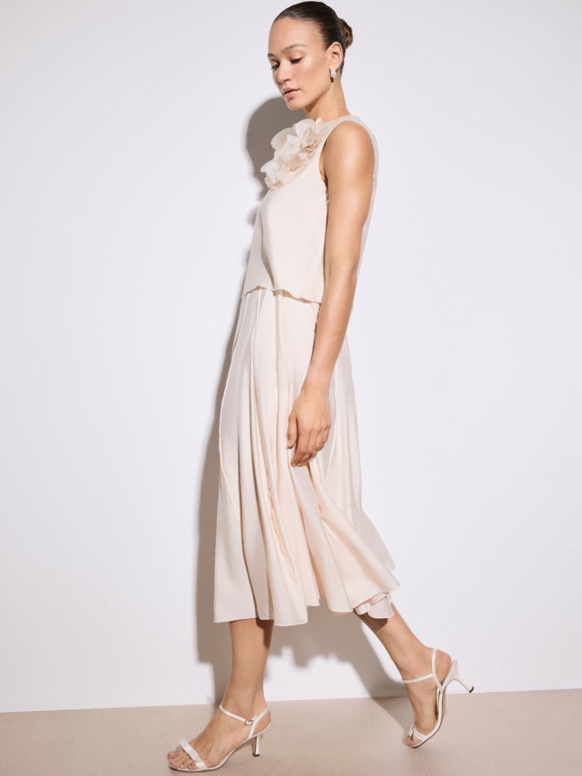 Detale eleganckiego kompletu damskiego: struktura plisowanej spódnicy w kolorze ecru oraz tiulowe kwiaty przy dekolcie topu, stylizacja z sandałami na pasku.