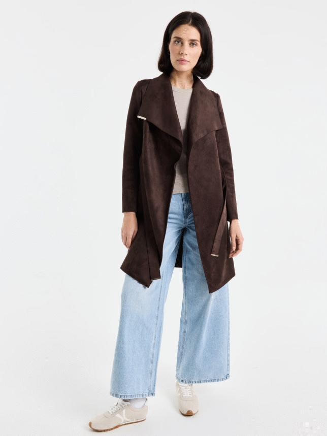 Kobieta w pełnej sylwetce, ubrana w rozpięty, ciemnobrązowy płaszcz z imitacji zamszu, jasny t-shirt oraz szerokie, błękitne jeansy i beżowe sneakersy.