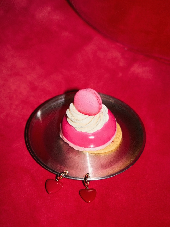 Różowe ciastko z makaronikiem na metalowym talerzyku obok kolczyków w kształcie czerwonych serc na intensywnie czerwonym tle.