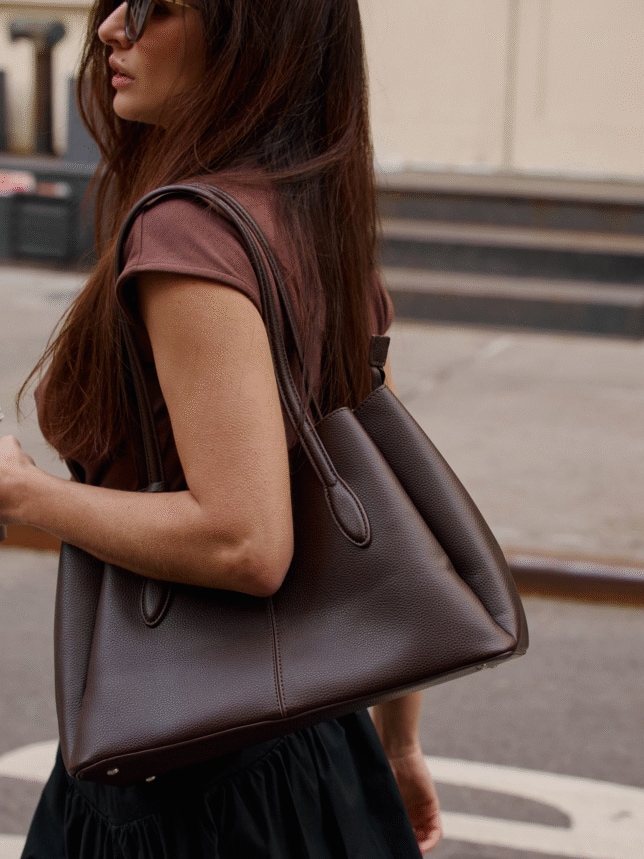 Zbliżenie na profil kobiety trzymającej na ramieniu dużą, strukturalną torebkę w kolorze gorzkiej czekolady. Kobieta ma na sobie ciemnobrązowy t-shirt i ciemną spódnicę, a na twarzy okulary przeciwsłoneczne. Tło miejskie z profilu.